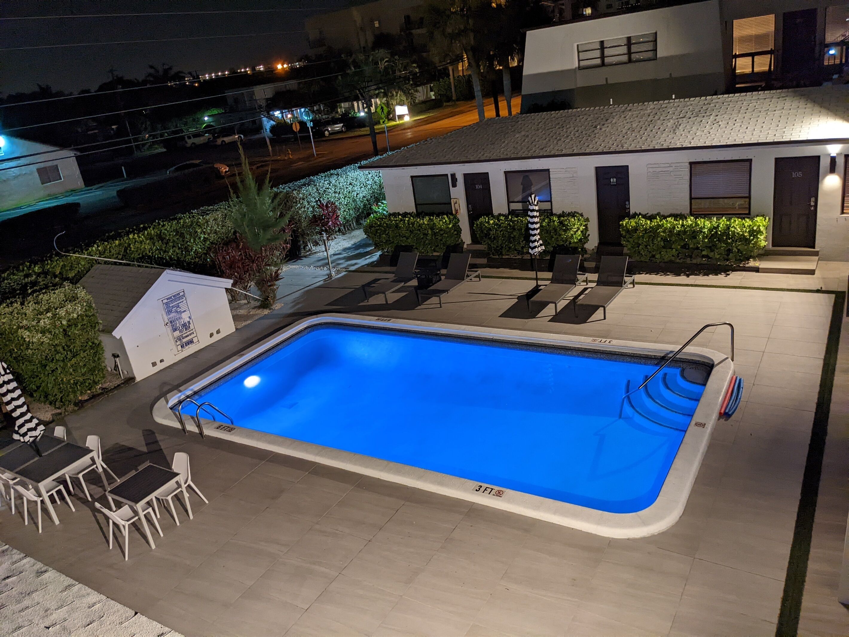 Uma piscina retangular com marcação de profundidade de 3 pés, rodeada por cadeiras de descanso e uma área de mesa pequena, com um edifício ao fundo.