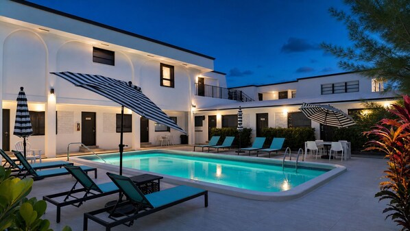 Una piscina con sillas de descanso y sombrillas, rodeada por un edificio con varias ventanas y puertas.