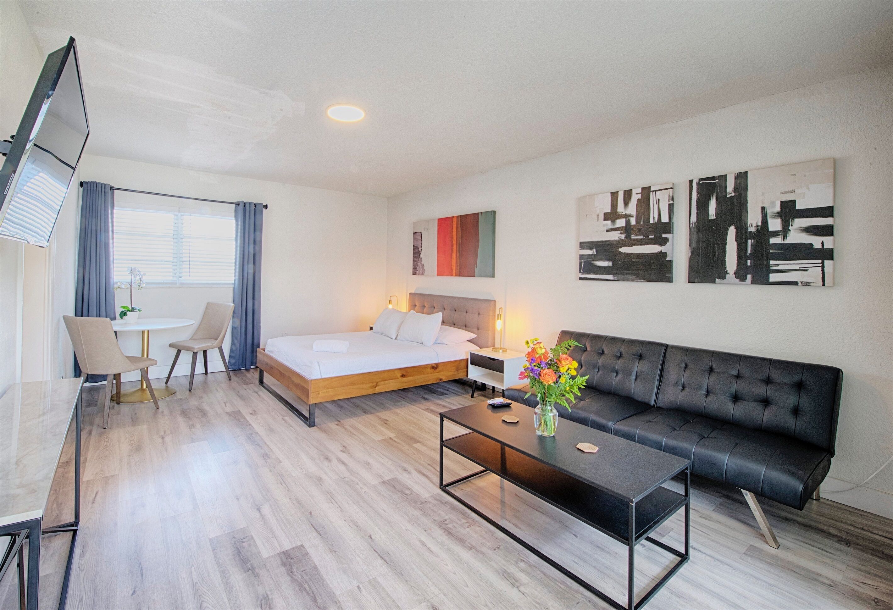 A modern hotel room with a bed, a sofa, a dining table with chairs, a TV, and artwork on the walls.