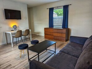 A modern living room with a sofa, a coffee table, a TV mounted on the wall, a desk with chairs, and a window with curtains. - Spacious 1BR #107 | Sleeps 4 | Beach Walk (Deerfield Beach)