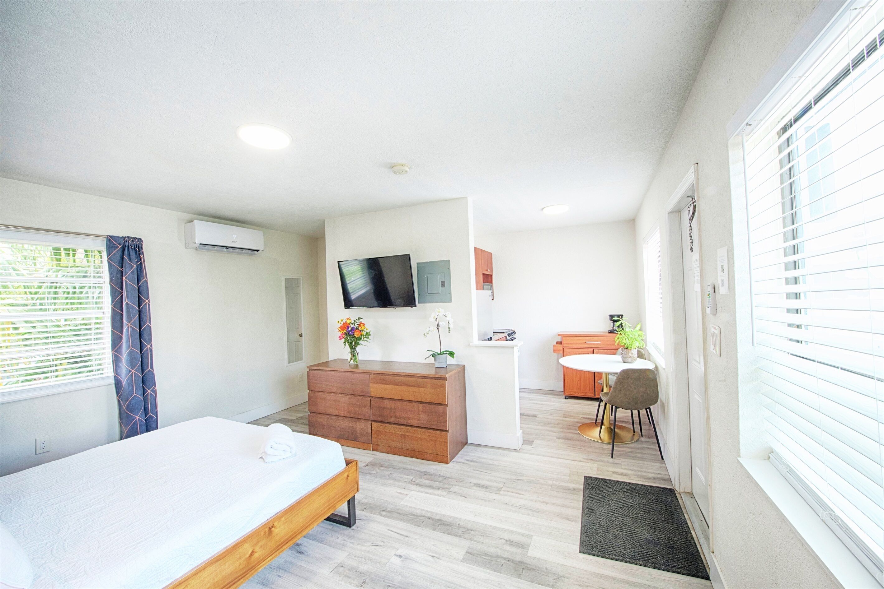 A modern hotel room with a bed, a wooden dresser, a small desk, a chair, and a TV mounted on the wall.