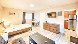 Ein modernes Hotelzimmer mit einem Bett, einem Schreibtisch, einem Esstisch, einer Couch und einer Küchenzeile mit einem Kühlschrank. An der Wand ist ein Fernseher angebracht.