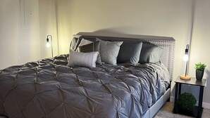 Une chambre avec un grand lit, une tête de lit, un tableau encadré au mur, une table de chevet avec une lampe et une petite plante.
