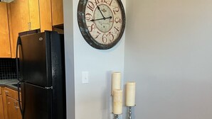 Une cuisine avec des meubles de rangement en bois, un réfrigérateur noir, une horloge murale et une petite table d’appoint avec des bougies.