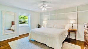 Ein Schlafzimmer mit Bett, Deckenventilator, Schminktisch und Fenster mit Blick auf Grünfläche.