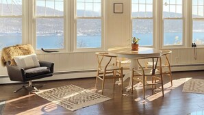 Una habitación con un ventanal grande que da a un cuerpo de agua y colinas, una mesa redonda con sillas, un plaid de piel en una silla y una alfombra estampada en el suelo.