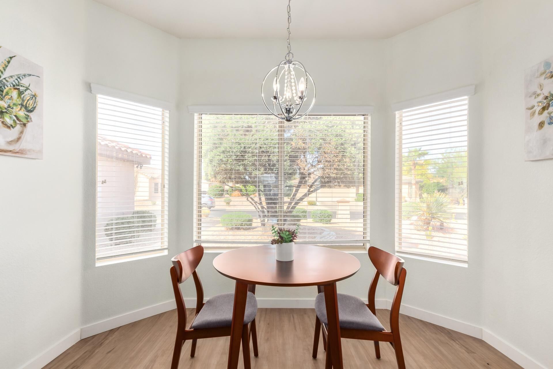 A dining area with a round table and four chairs, a chandelier, and large windows with blinds.