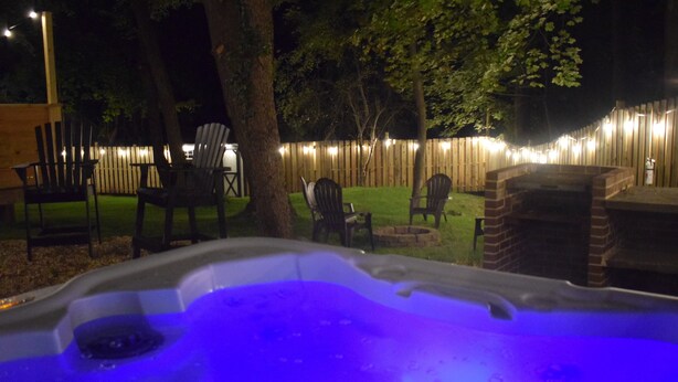 Un jacuzzi con borde de ladrillo, rodeado de sillas de madera y una valla de madera con luces colgantes.