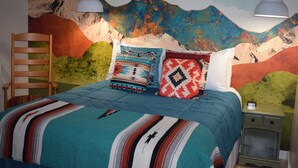 Un dormitorio con un mural de montañas, una cama con ropa de cama estampada, dos lámparas colgantes, una silla de madera y una mesita de noche.