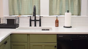 Una cocina con armarios verdes, una encimera blanca, un grifo negro, un escurreplatos, una botella de jabón y un dispensador de toallas de papel.