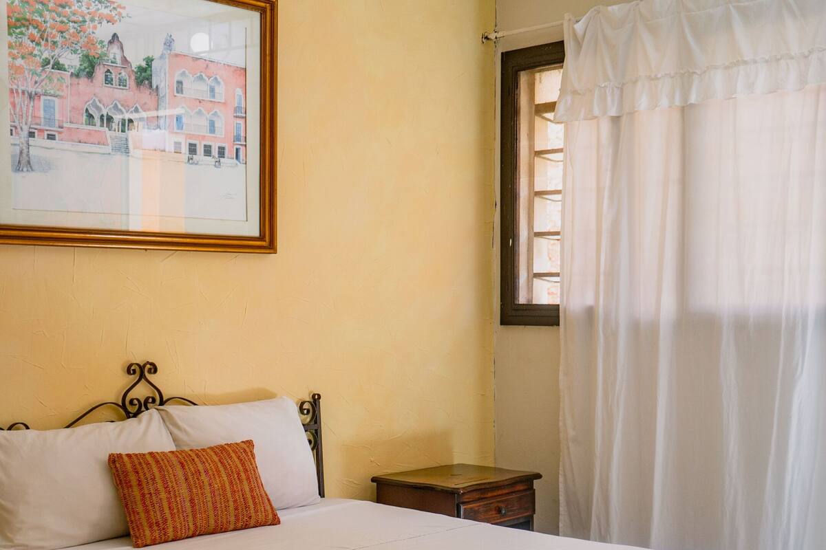 A bedroom with a bed, a ceiling fan, a nightstand, a window with curtains, and a painting on the wall.