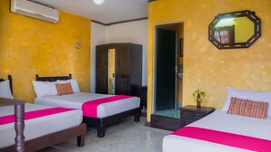 Quarto de hotel com três camas, cada uma com colcha rosa e travesseiros brancos. Possui um guarda-roupa de madeira, um espelho na parede e uma pequena planta em uma mesa de cabeceira.