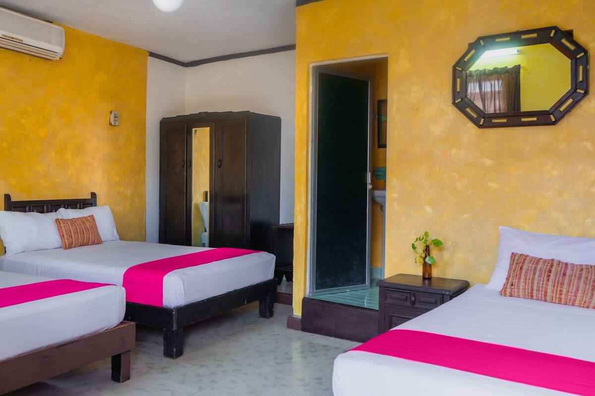 A hotel room with three beds, each with a pink bedspread and white pillows. There is a wooden wardrobe, a mirror on the wall, and a small plant on a nightstand.