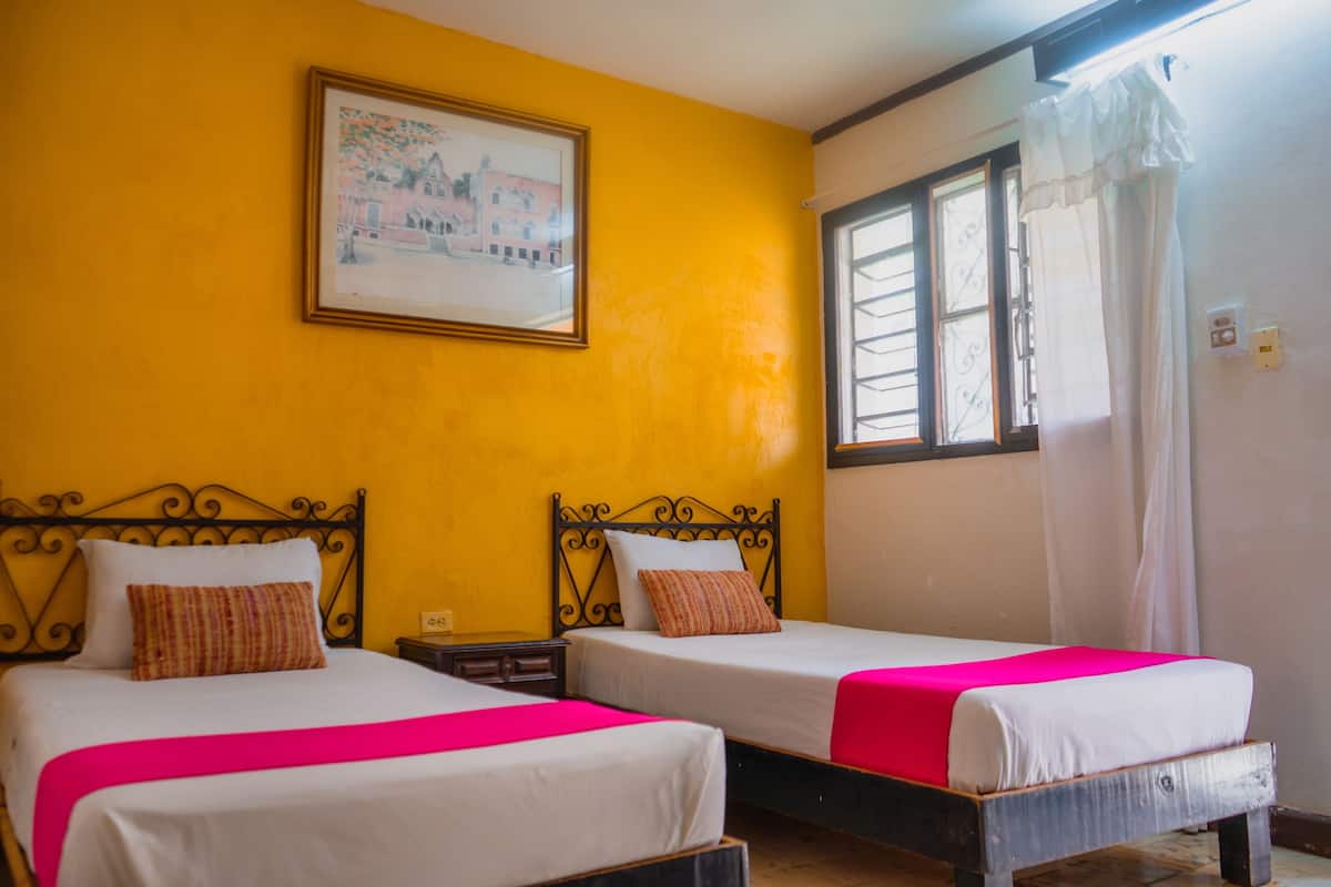A room with two beds, yellow walls, a framed picture, and a window with curtains.