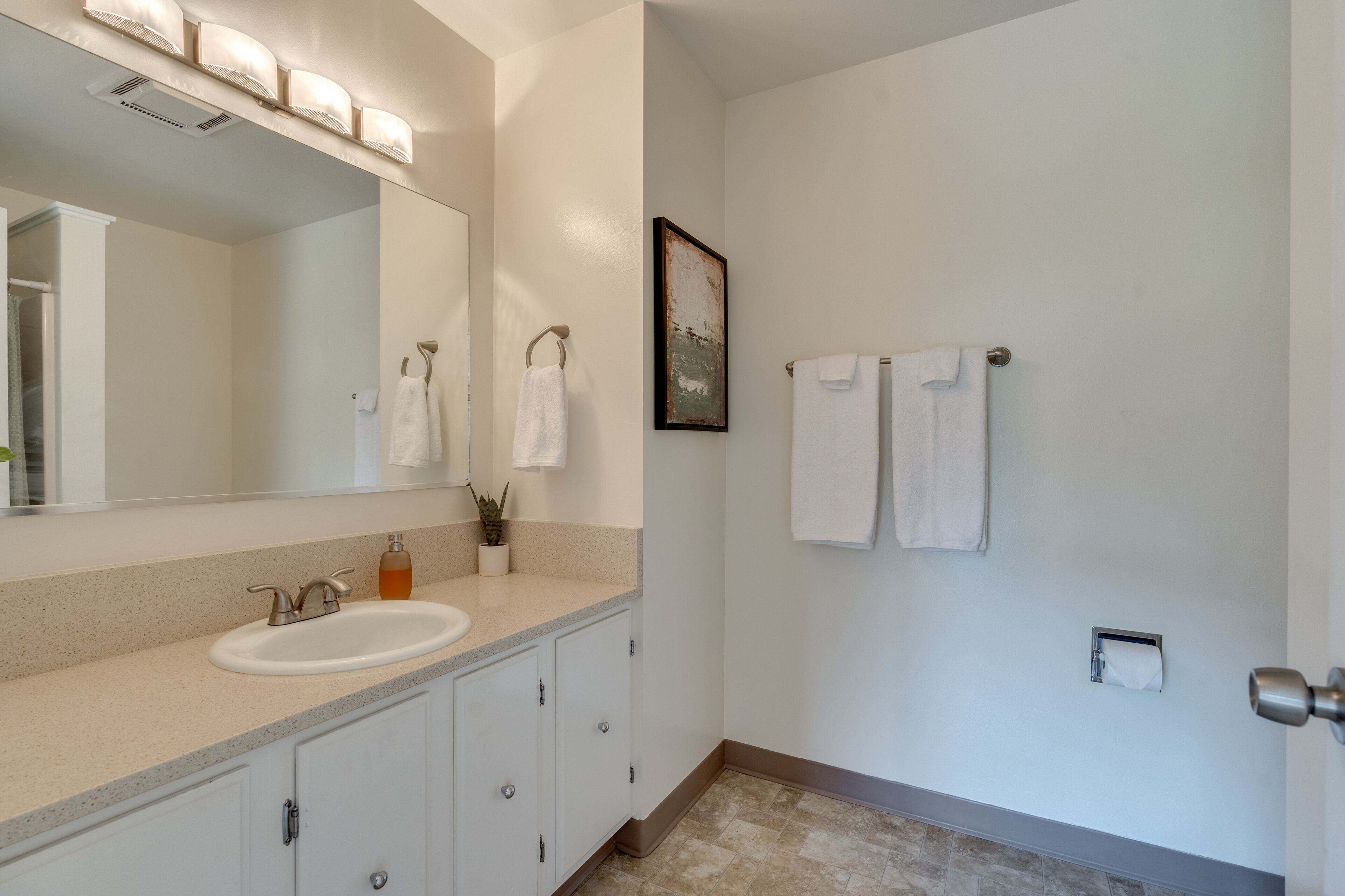Un baño moderno con un lavamanos y tocador blancos, y un espejo grande. Hay dos toallas colgadas en la pared, y una pequeña planta en maceta sobre la encimera.