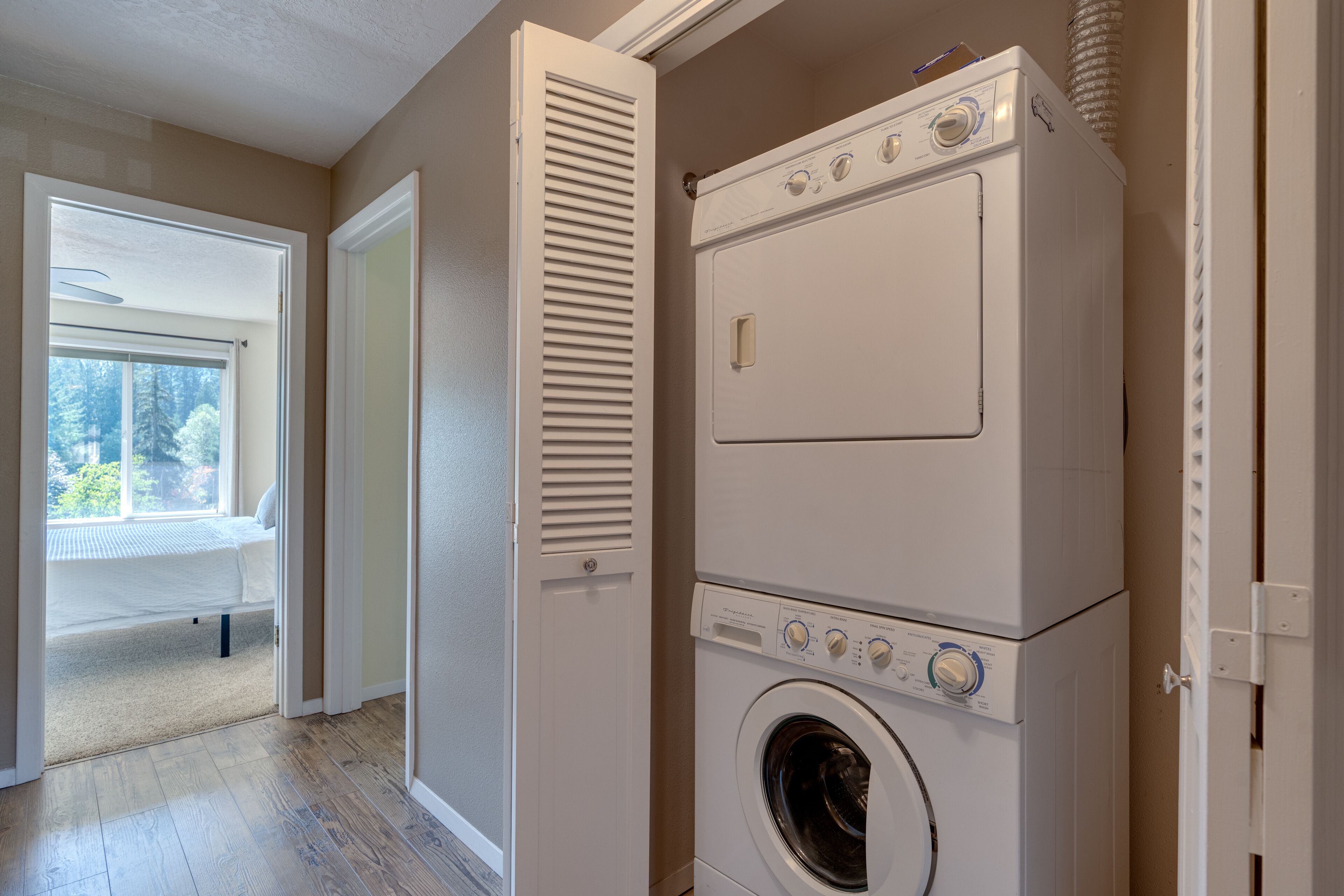 A laundry room with a stacked washer and dryer, a door leading to a bedroom, and a window with a view of trees.