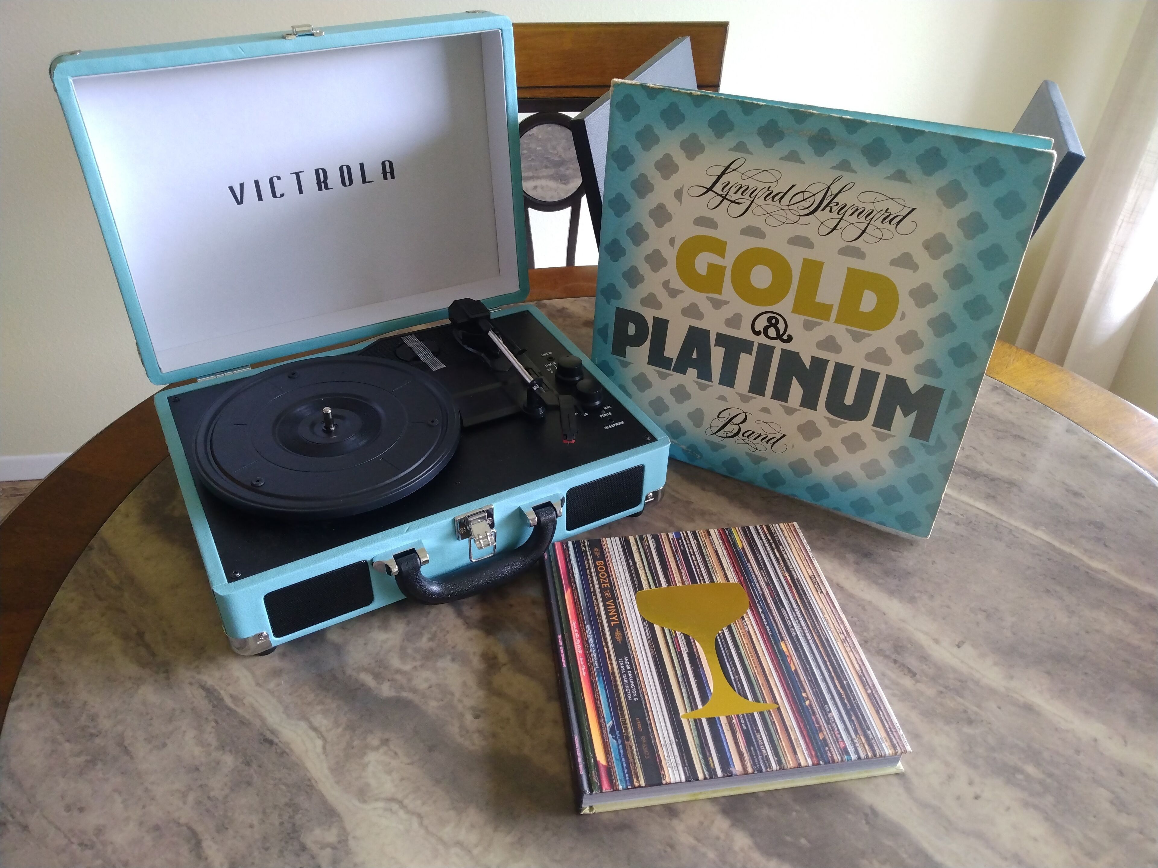 Tocadiscos de vinilo en una funda azul claro, con un disco de vinilo adentro, una caja de discos con la etiqueta 'Gold & Platinum Band' y una caja de discos con un diseño de copa de vino dorada.
