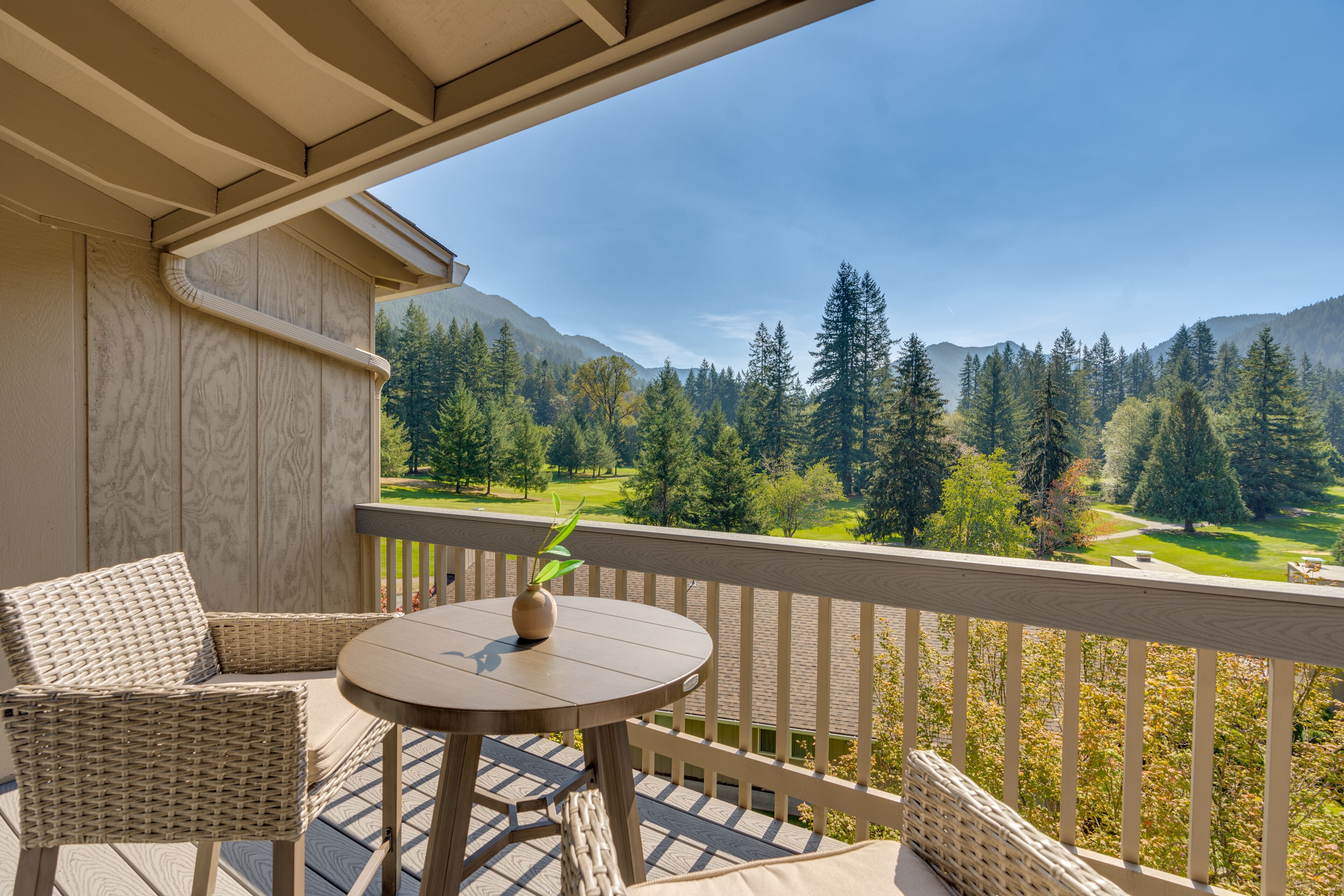 Un balcon doté de meubles en osier et d’une table, offrant une vue sur une zone boisée comprenant un terrain de golf et des montagnes.