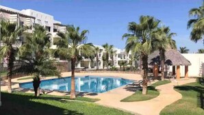 A pool area with palm trees, a thatched-roof hut, and a white building with balconies. - Apartament 2 beds+pool+gym+ petfriendly (Cabo San Lucas)