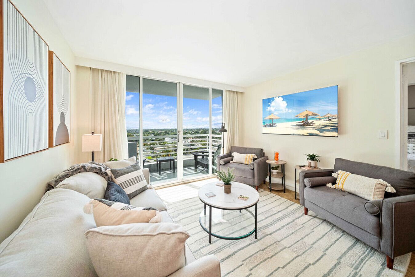 A spacious living room with a large sofa, a smaller sofa, a coffee table, and a balcony with a view of the city.