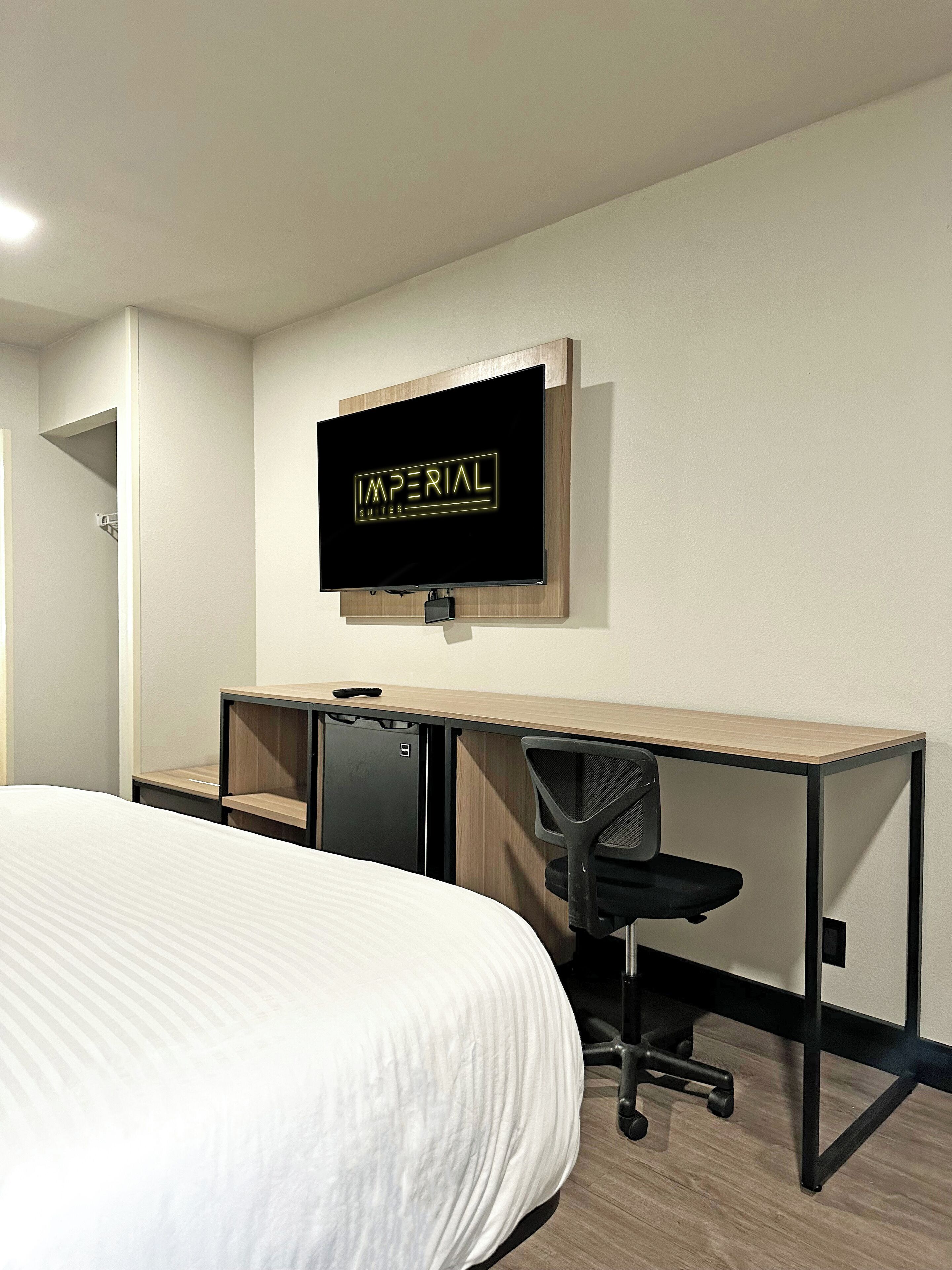 Ein Hotelzimmer mit einem Bett, einem Schreibtisch mit Computer, einem Sessel und einem an der Wand befestigten Fernseher, der "IMPERIAL SUITES" anzeigt.