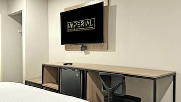 Ein Hotelzimmer mit einem Bett, einem Schreibtisch mit Computer, einem Sessel und einem an der Wand befestigten Fernseher, der "IMPERIAL SUITES" anzeigt.