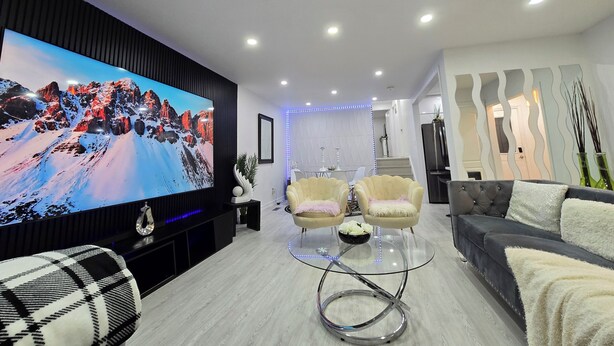 A modern living room with a large flat-screen TV, two armchairs, a sofa, and a glass coffee table. - Ultimate Retreat: 98" Cinema + Arcade Game Room + BBQ + Airport (Brampton)