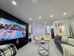 A modern living room with a large flat-screen TV, two armchairs, a sofa, and a glass coffee table. - Ultimate Retreat: 98" Cinema + Arcade Game Room + BBQ + Airport (Brampton)