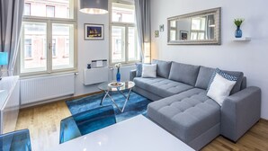 Un soggiorno moderno con un divano ad angolo grigio, un tavolino in vetro e un tappeto blu. Si vedono due finestre con tende, un termosifone bianco e uno specchio fissato al muro.