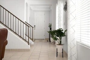 A hallway with a staircase, a white door at the end, and potted plants along the wall. - Stylish Central Tucson Family-sized Home with Bikes to Ride (Tucson)