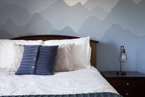 Uma cama bem arrumada com um edredom branco e duas almofadas, uma azul e uma branca, em um quarto com papel de parede com tema de montanhas.