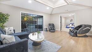 A spacious living room with a high ceiling, a large window, a sofa, a massage chair, and a small table with a vase. - ★4BR Dallas Family Estate | Heated Pool + Hot Tub | Sleeps 16 | Lake Highlands★ (Dallas)