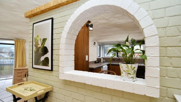 Una habitación con pared de ladrillo blanco, un armario de madera, un cuadro enmarcado, una planta en maceta y una ventana con vistas.