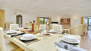 Un comedor con una larga mesa dispuesta para una comida, incluyendo platos, vasos y salero y pimentero. Hay sillas alrededor de la mesa y se ve una estantería al fondo.