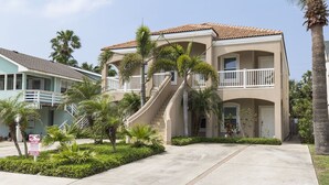 A two-story building with a beige facade, a tiled roof, and a central staircase. There are palm trees and a well-maintained lawn in front. - Cushy Crab, Pool and 3 Minute Walk to Beach!! (South Padre Island)