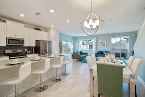 A modern kitchen with white cabinets, a built-in microwave, and a stainless steel refrigerator. There is a dining area with a white table and chairs, and a large window providing natural light. - Champions Gate by CozyKey (Davenport)