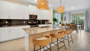 A modern kitchen with white cabinets, a central island with bar stools, and pendant lighting. - Champions Gate by CozyKey (Davenport)