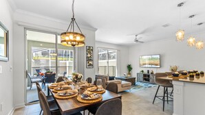 A spacious dining area with a long wooden table set for a meal, a chandelier, and a view of the outdoors through large windows. - Champions Gate by CozyKey (Davenport)