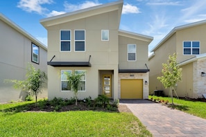 A modern two-story house with a yellow garage door, a small front yard with young trees, and a brick pathway leading to the entrance. - Champions Gate by CozyKey (Davenport)