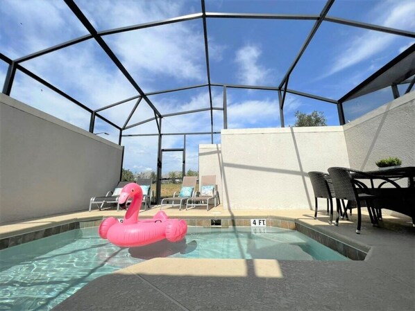 A covered outdoor swimming pool with a pink flamingo float, lounge chairs, and a table. - Champions Gate by CozyKey (Davenport)