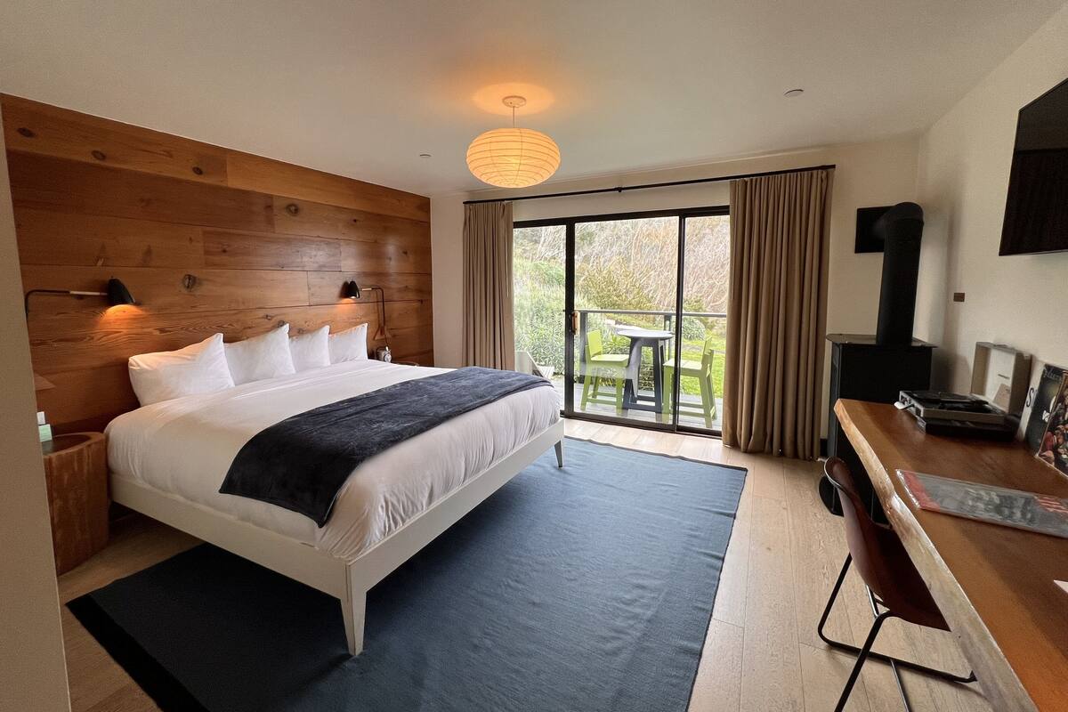 Quarto com cama grande, cabeceira de madeira, mesa de cabeceira, abajur e vista para uma varanda com mobiliário.