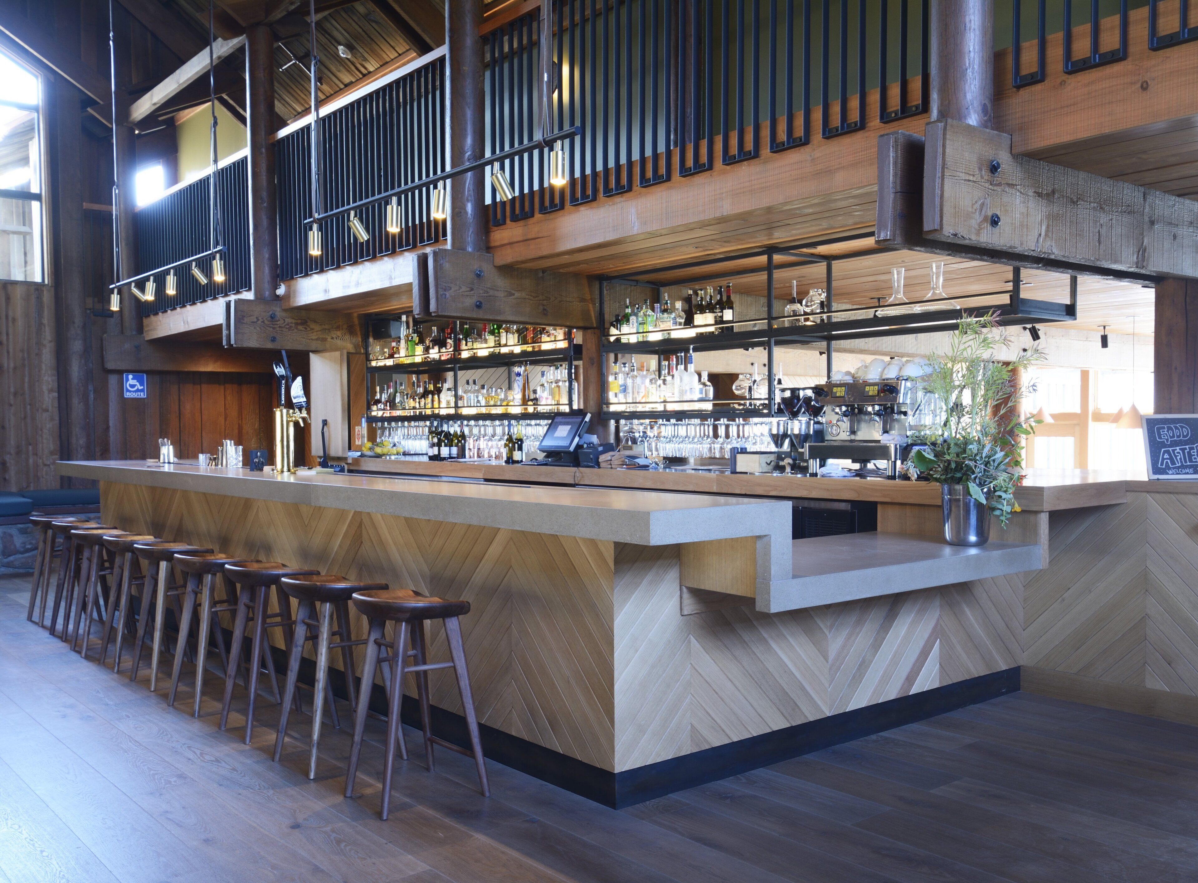 Un bar con mostrador de madera, taburetes y una variedad de botellas y vasos.