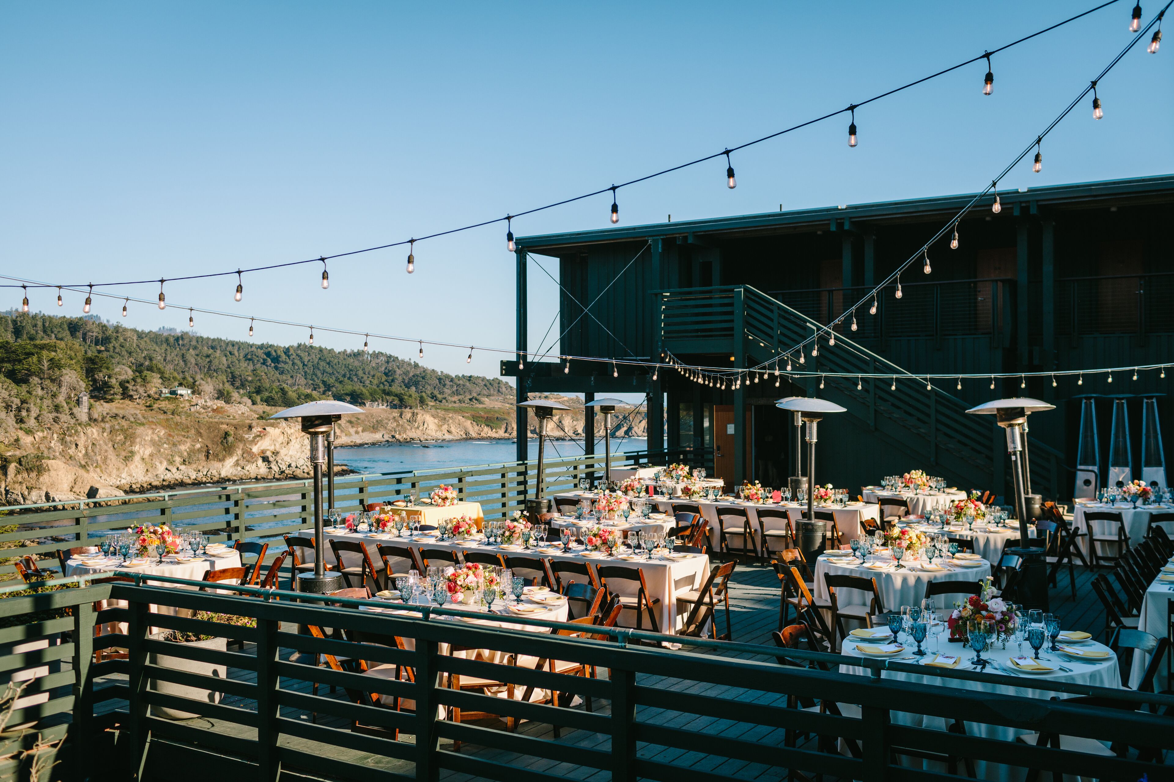 An outdoor dining area with tables set for a meal, overlooking a body of water and a forested hillside.