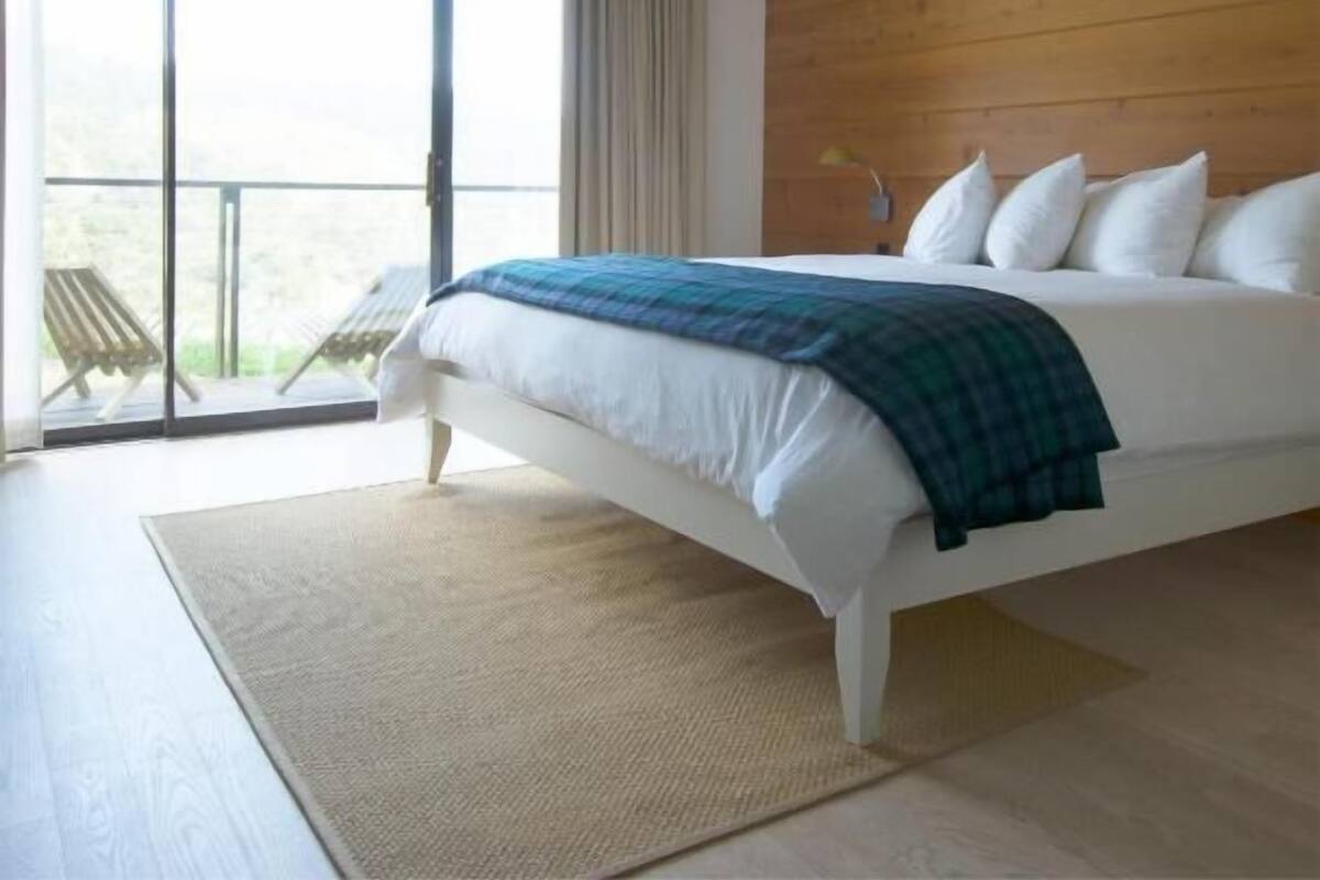 Quarto moderno com cama grande, cabeceira de madeira, varanda com cadeiras e vista para o exterior.