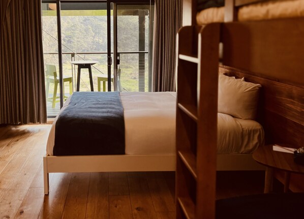 A wooden bed with a mattress and pillows, a wooden floor, a window with curtains, and a view of greenery outside.
