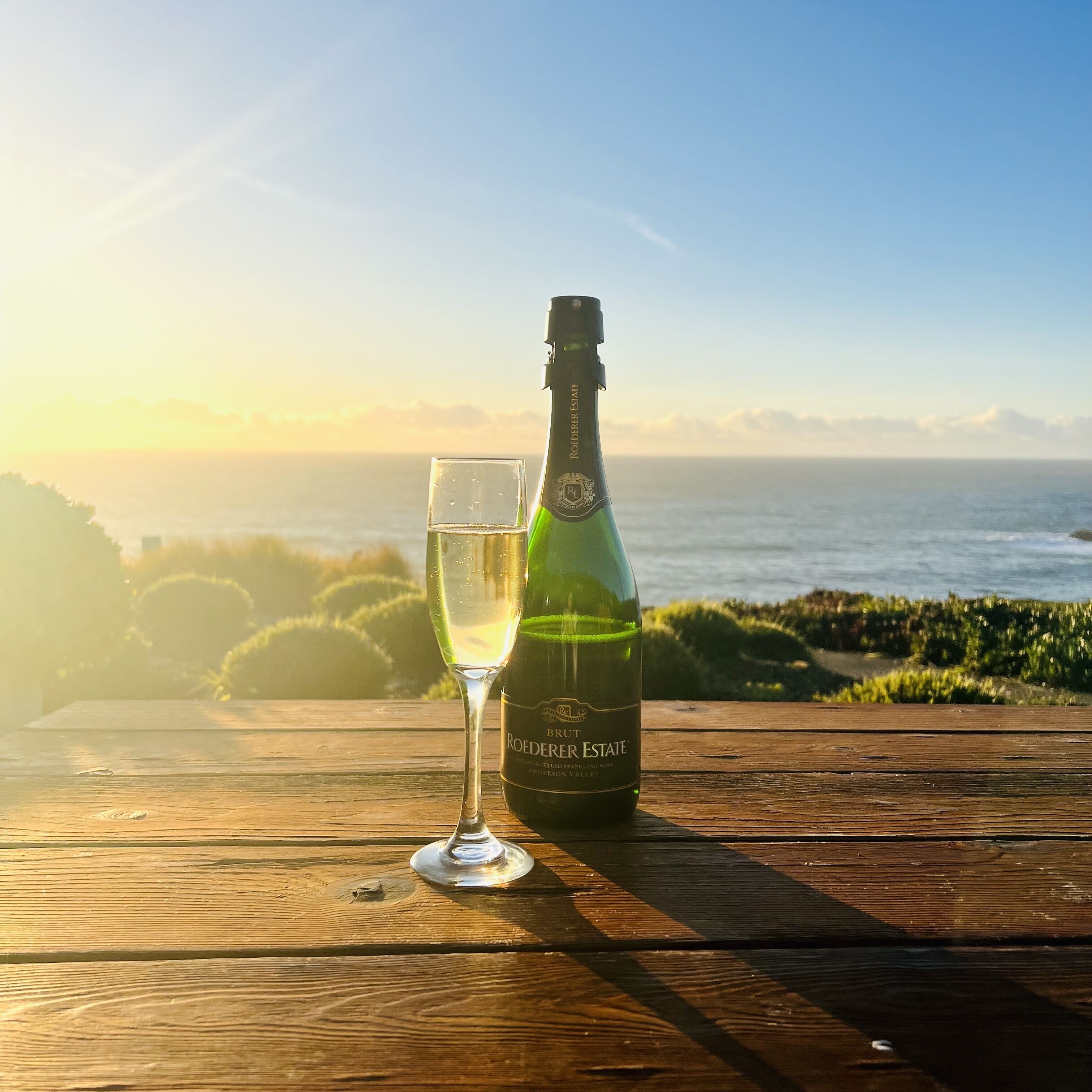 A bottle of Roederer Estate Brut champagne and a glass on a wooden table with a view of the ocean and hills in the background.