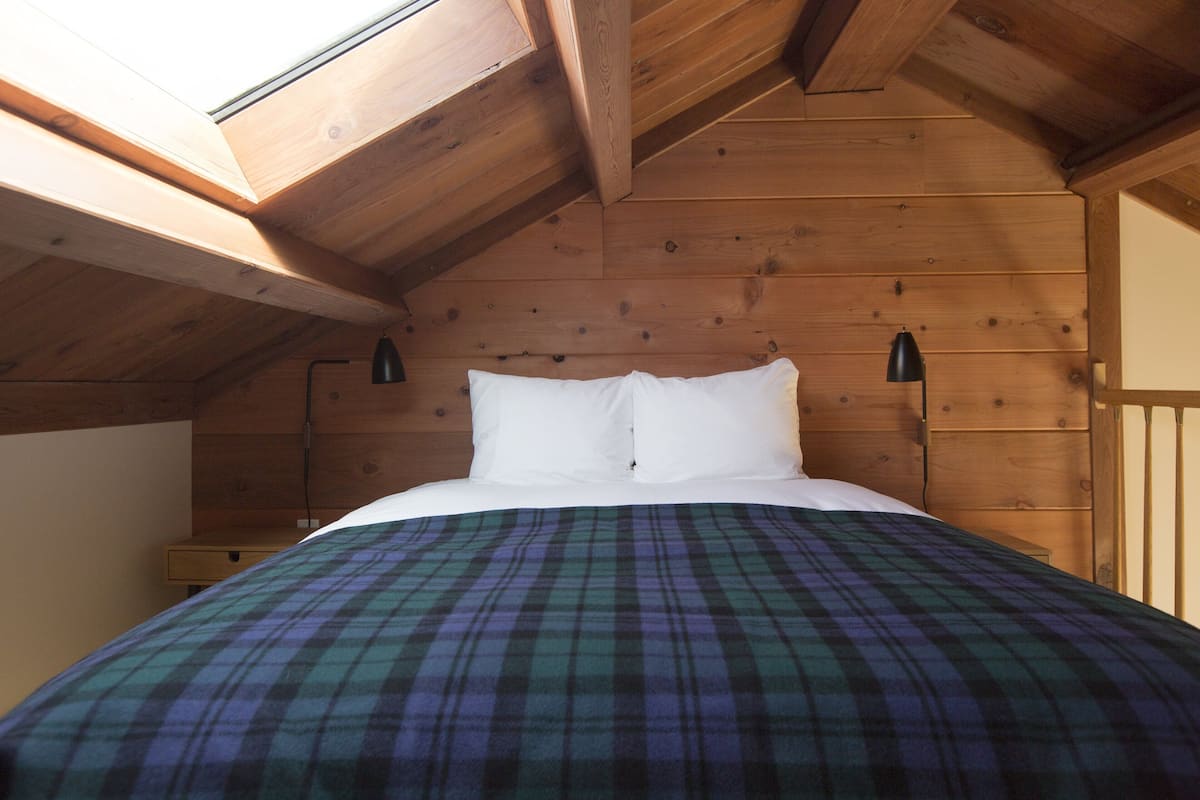 Quarto com teto de madeira, cama com roupa de cama branca e manta xadrez.