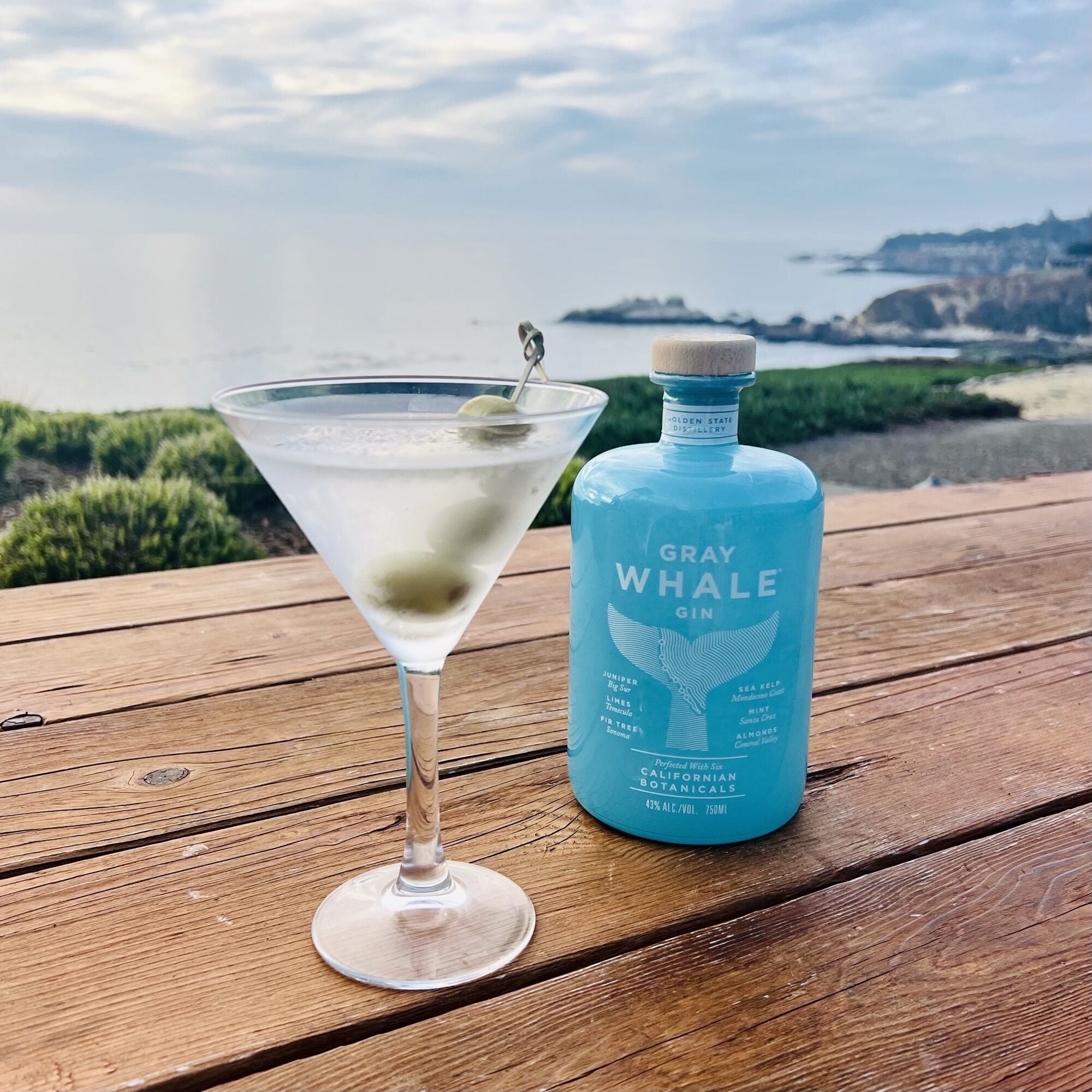 Ein Martini-Glas, gefüllt mit klarer Flüssigkeit und einer Limettenscheibe, steht auf einem Holztisch neben einer Flasche Gray Whale Gin.