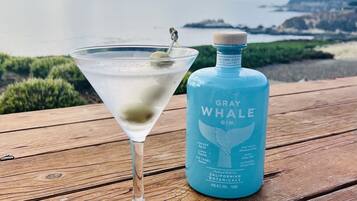 Una copa de martini llena de un líquido transparente y una rodaja de lima, sobre una mesa de madera junto a una botella de Gray Whale Gin.