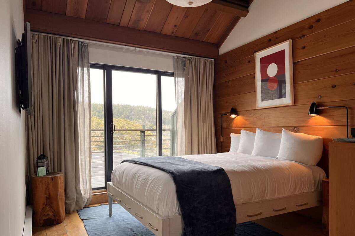 Quarto com uma cama grande, paredes de madeira, ventilador de teto, mesa de cabeceira e vista para o exterior.